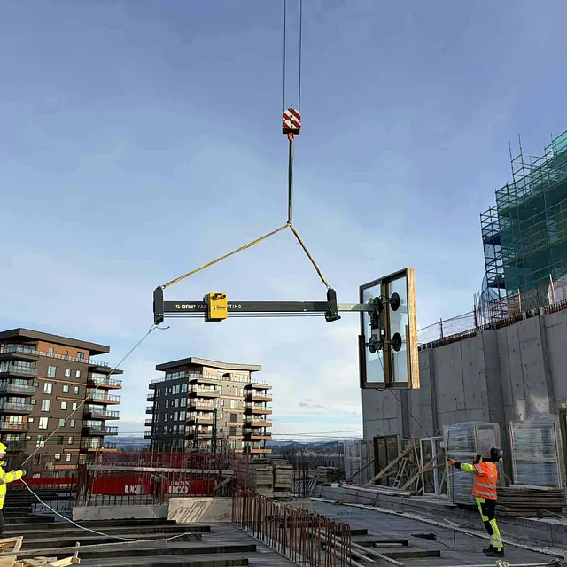 Stor glassmodul løftes med kran og vakuumløfter på byggeplass med arbeidere som styrer løftet.