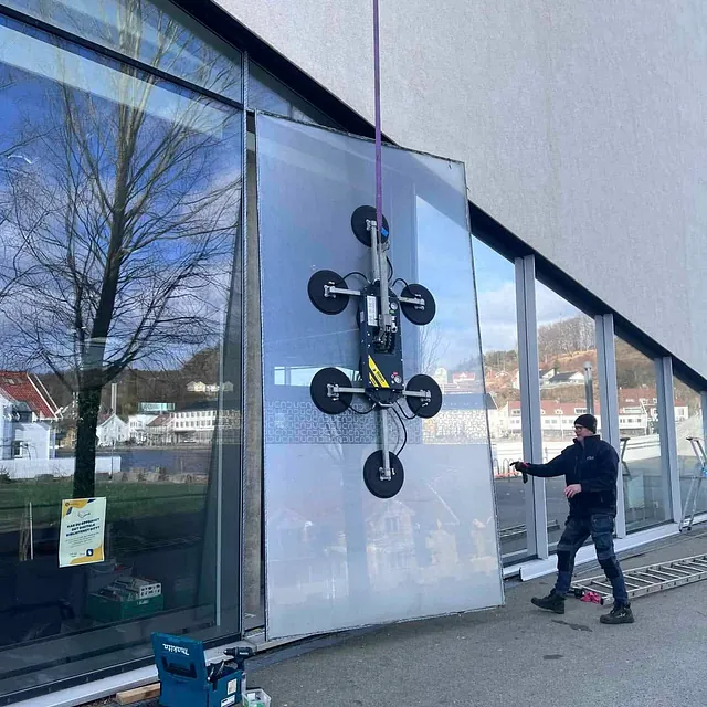 Arbeider styrer stor glassplate med vakuumløfter foran moderne bygg med glassfasade.