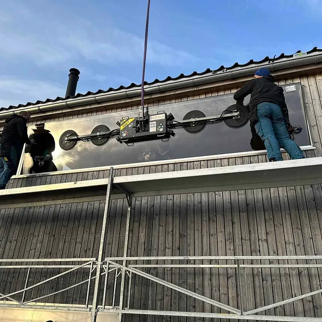 To arbeidere monterer lang horisontal glassrute med vakuumløfter på trebygning.