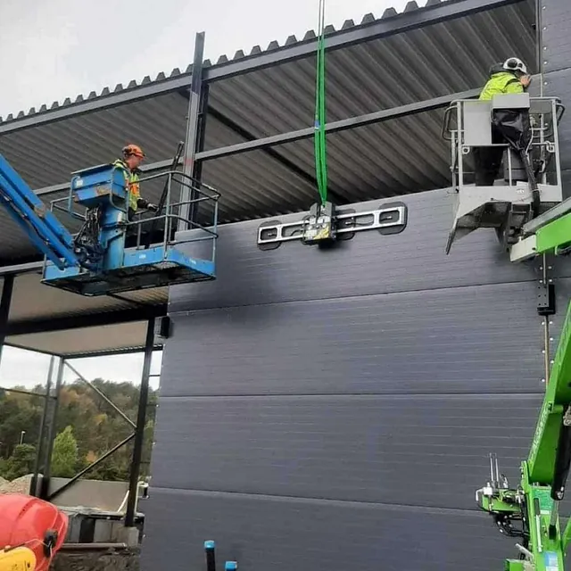 Arbeidere monterer fasadeelement med vakuumløfter ved hjelp av lift og kran på industribygg.