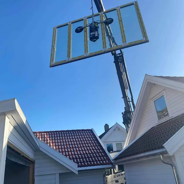 Stor glassfasade løftes med kran og vakuumløfter over hustak i boligområde.