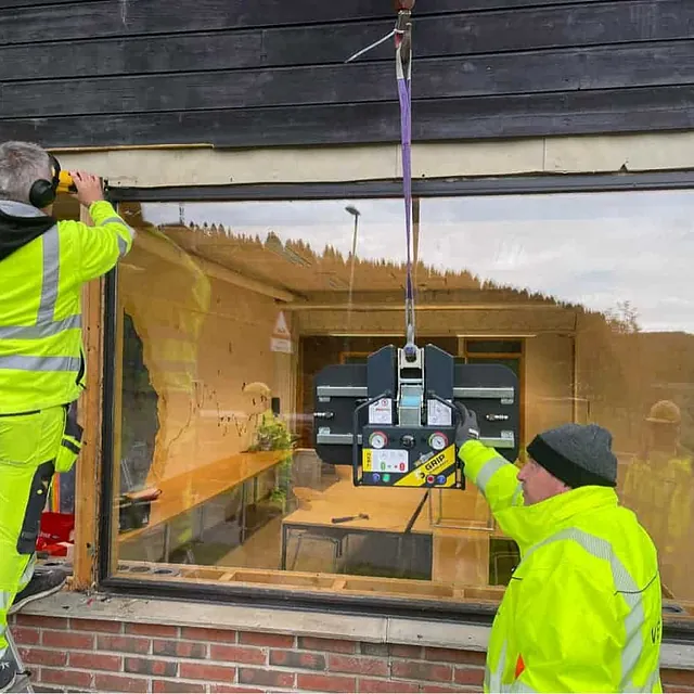 To arbeidere monterer stor glassrute med vakuumløfter i vegg på bolig.